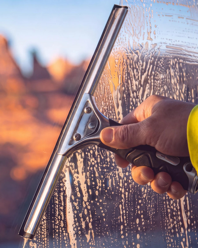 A window cleaner's hand pulling a squeegee through soapy water on a window, red rock cliffs blurred beyond
